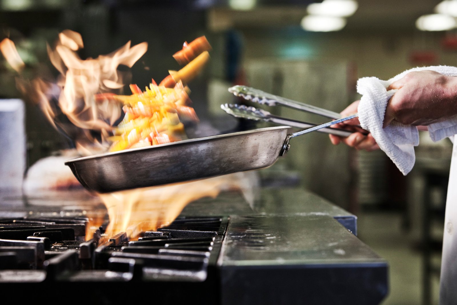 Close-up of chef's hands holding a sauté pan to cook food, flambeing contents. Flames rising from the pan.