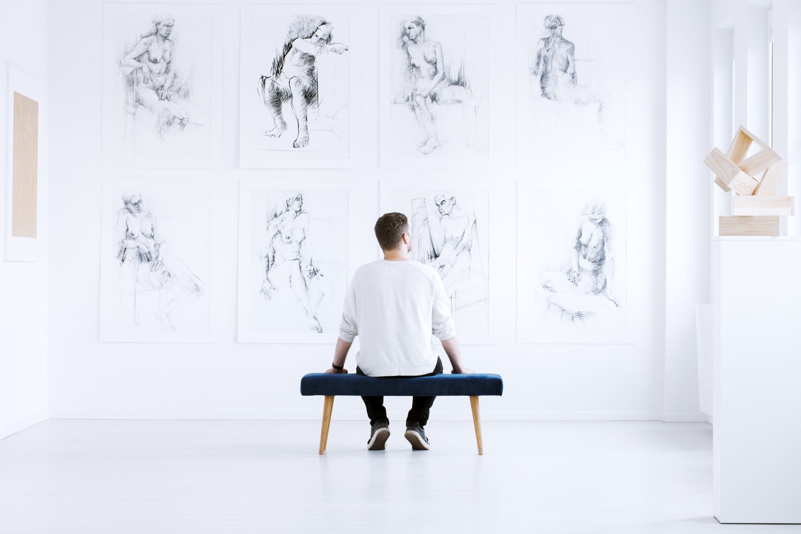 Man relaxing in art gallery while sitting on stool in front of white wall with drawings. Art gallery concept